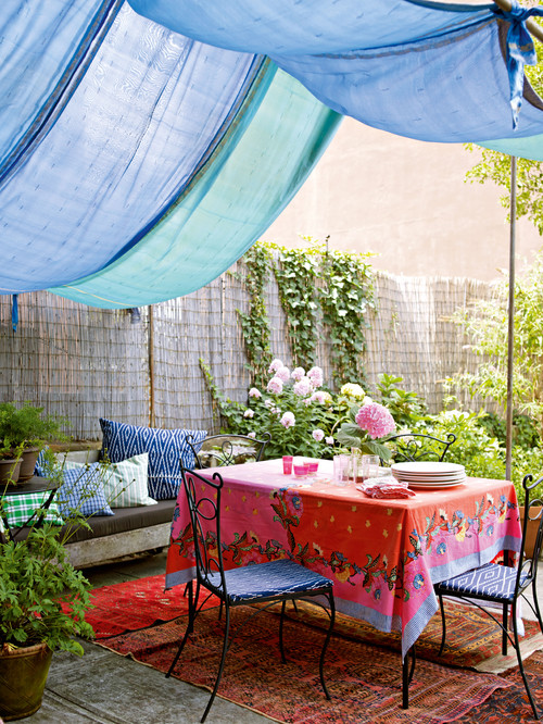 Outdoor patio with blue draped fabric canopy, colorful red and pink floral tablecloth, wrought iron chairs, and lush greenery